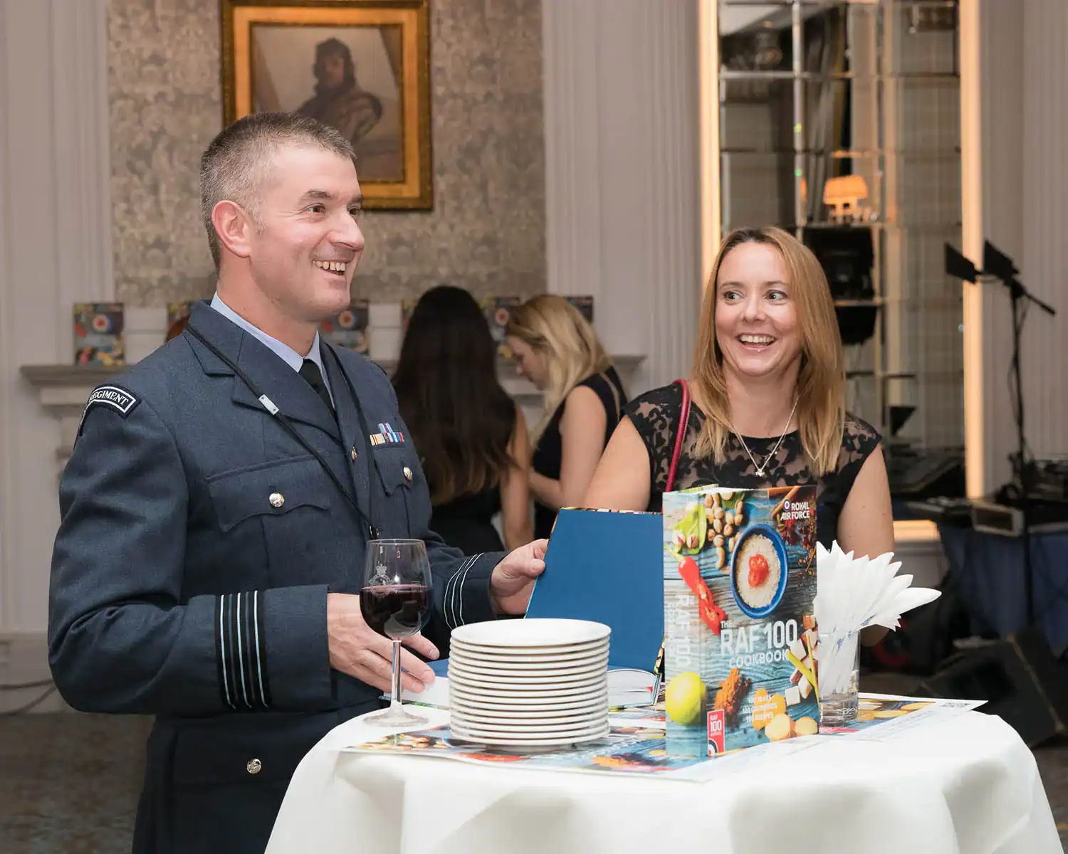 A man in a military uniform and a woman stand by a table adorned with plates and the "RAF 100 Cookbook" in a beautifully decorated room.