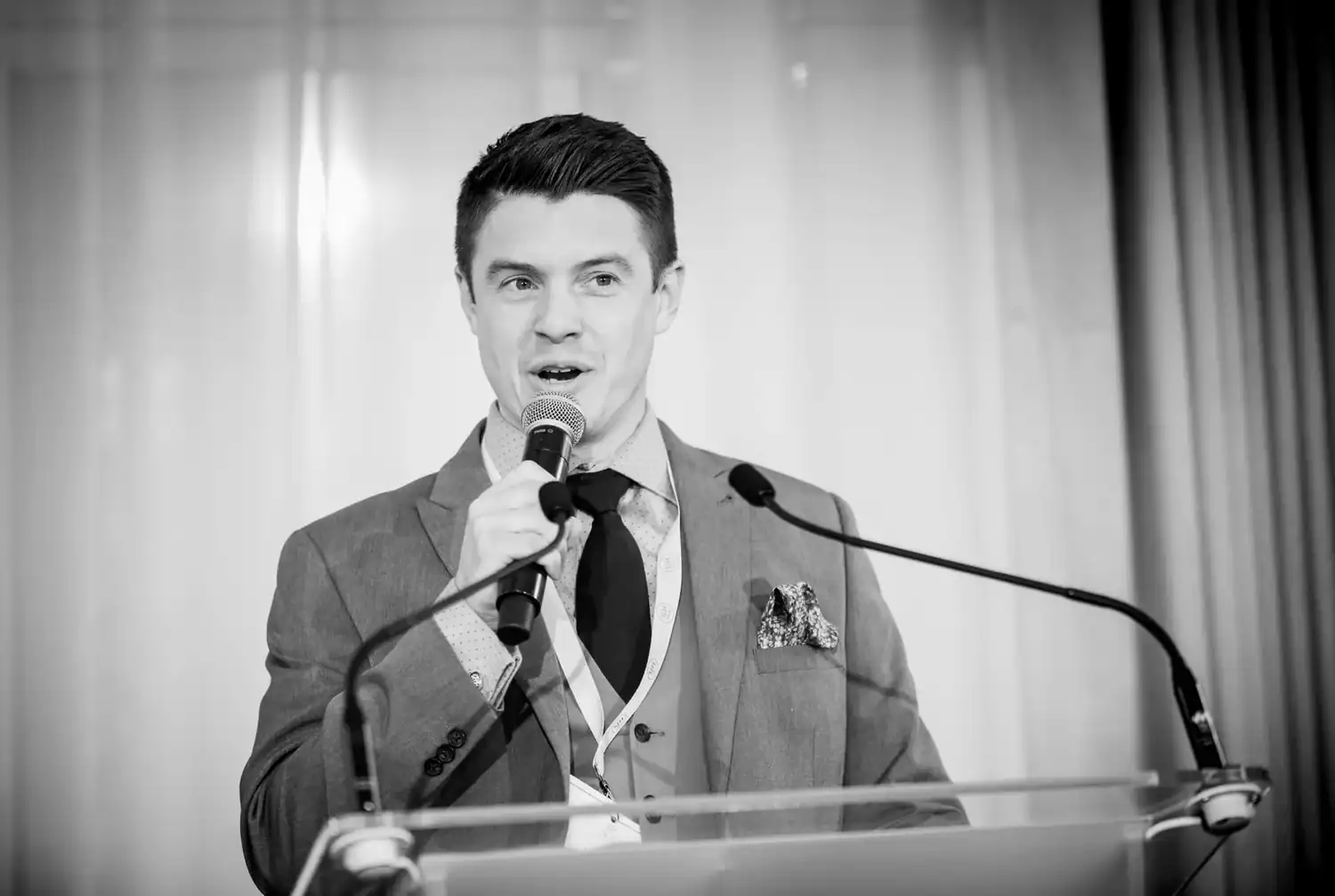 A man in a suit and tie speaks into a microphone at a podium, with curtains in the background, channeling the quiet strength of everyday heroes. Black and white image.