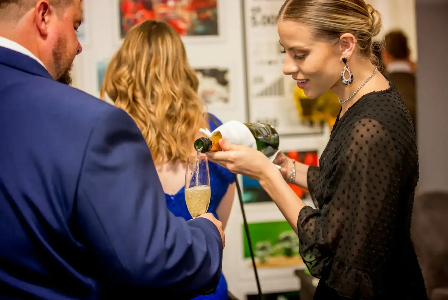 At a lively gathering, a woman, akin to the heroes of hospitality, gracefully pours champagne into a glass held by a man in a blue suit.