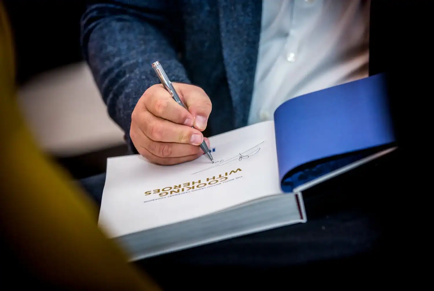 A person in a blue jacket and white shirt signs a book titled "COOKING WITH HEROES," deftly combining their passion for cooking with admiration for heroes.