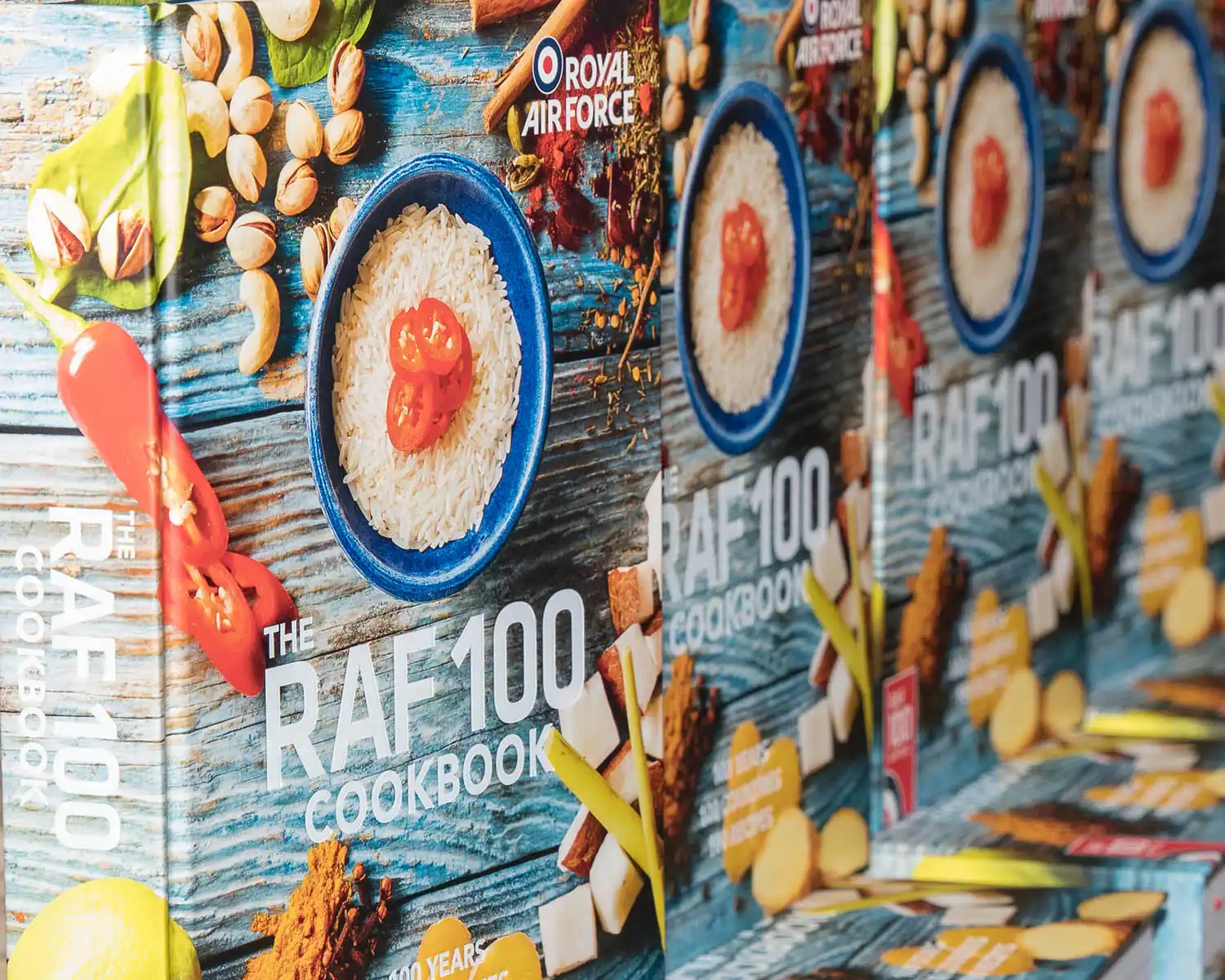Rows of "The RAF 100 Cookbook" proudly showcase its cover, featuring a bowl of white rice garnished with red peppers, surrounded by various ingredients and the distinctive Royal Air Force branding.