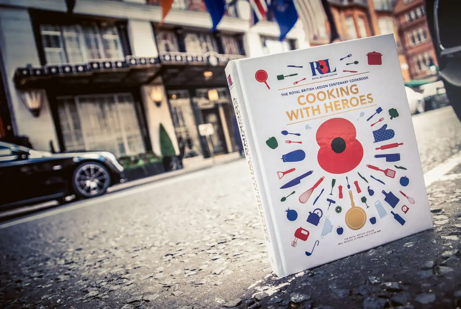 A cookbook titled "Cooking with Heroes" is artfully displayed on a street in front of an ornate building, its cover surrounded by whimsical illustrations of kitchen utensils.