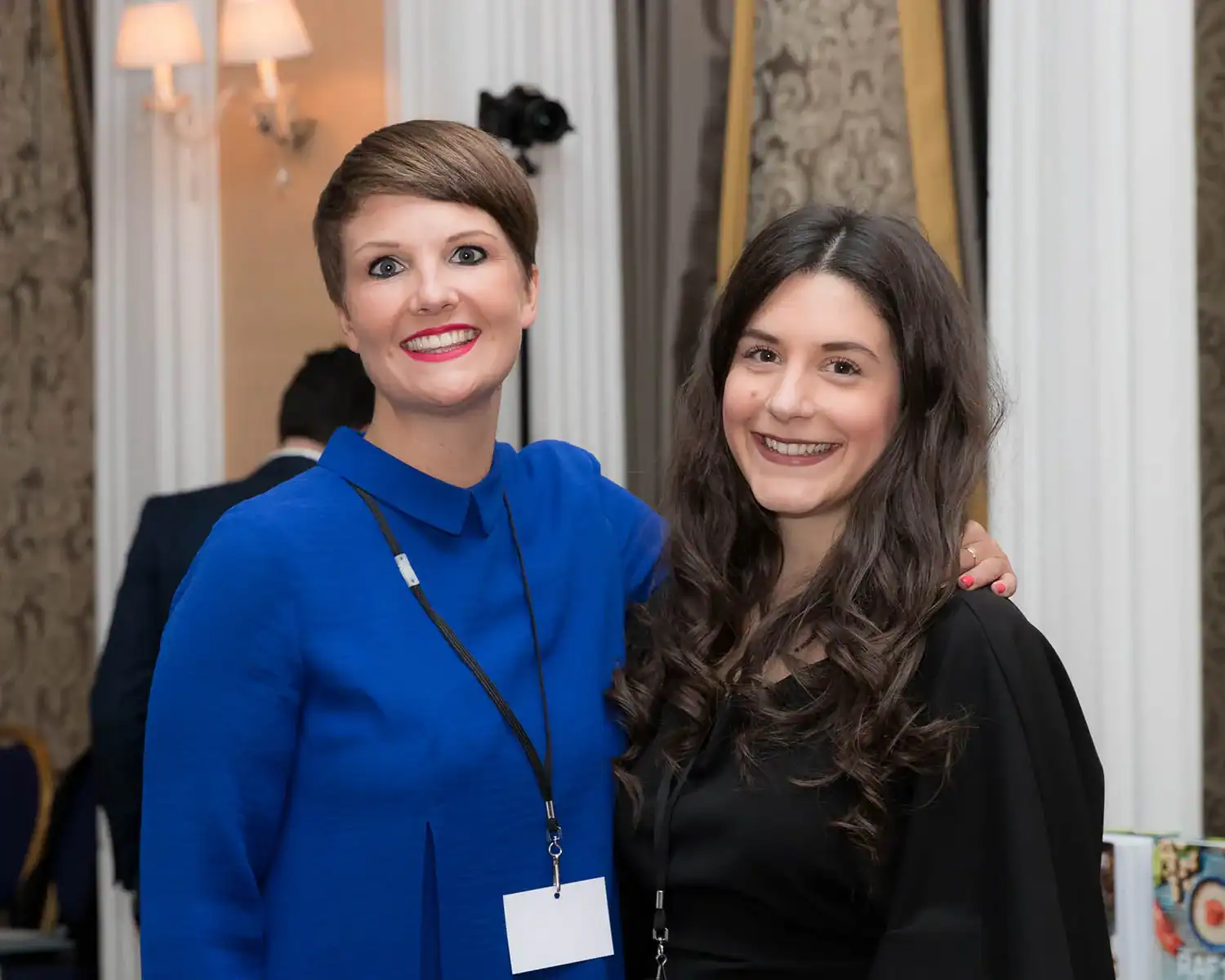Two women with conference badges smile warmly, one in a blue outfit and the other in black, standing indoors as they discuss the RAF 100 Cookbook.