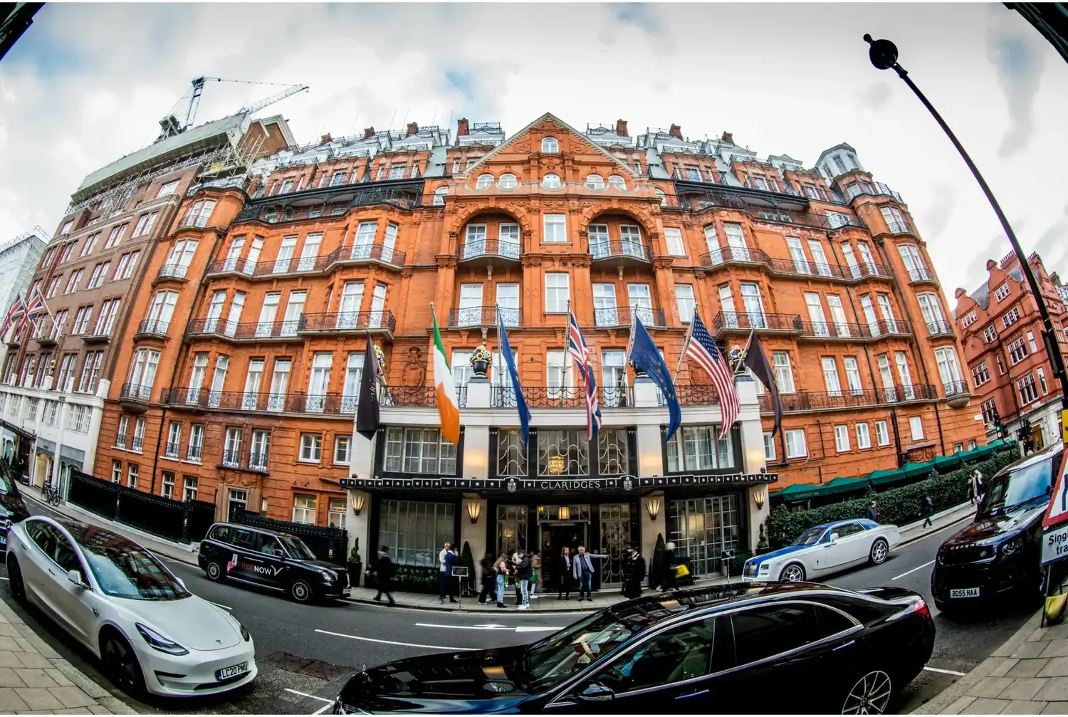 Street view of a large, ornate red brick building with flags at the entrance, reminiscent of a place where everyday heroes might gather. Several cars and people are visible in front, along with a lamppost in the foreground.