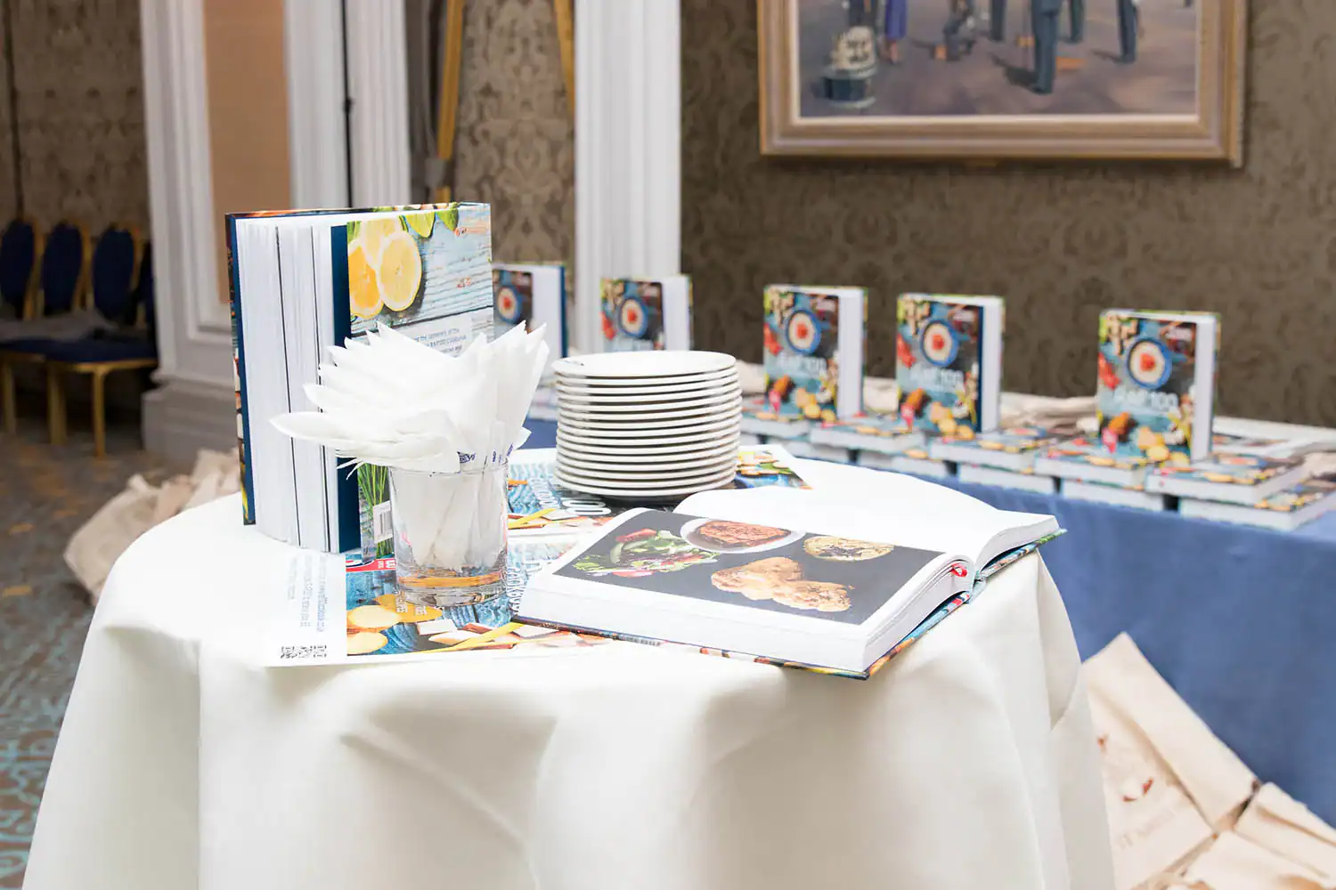A table with stacked books, an RAF 100-themed open cookbook, white plates, and neatly folded napkins. More books are displayed on a table in the background.