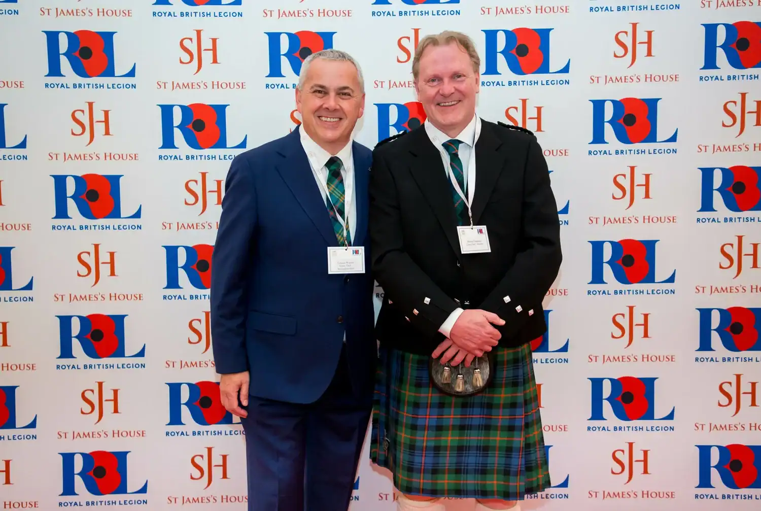 Two men stand smiling like heroes on a red carpet in front of a backdrop with logos from the Royal British Legion and St. James's House. One wears a suit, the other a traditional Scottish kilt outfit, both embodying elegance.