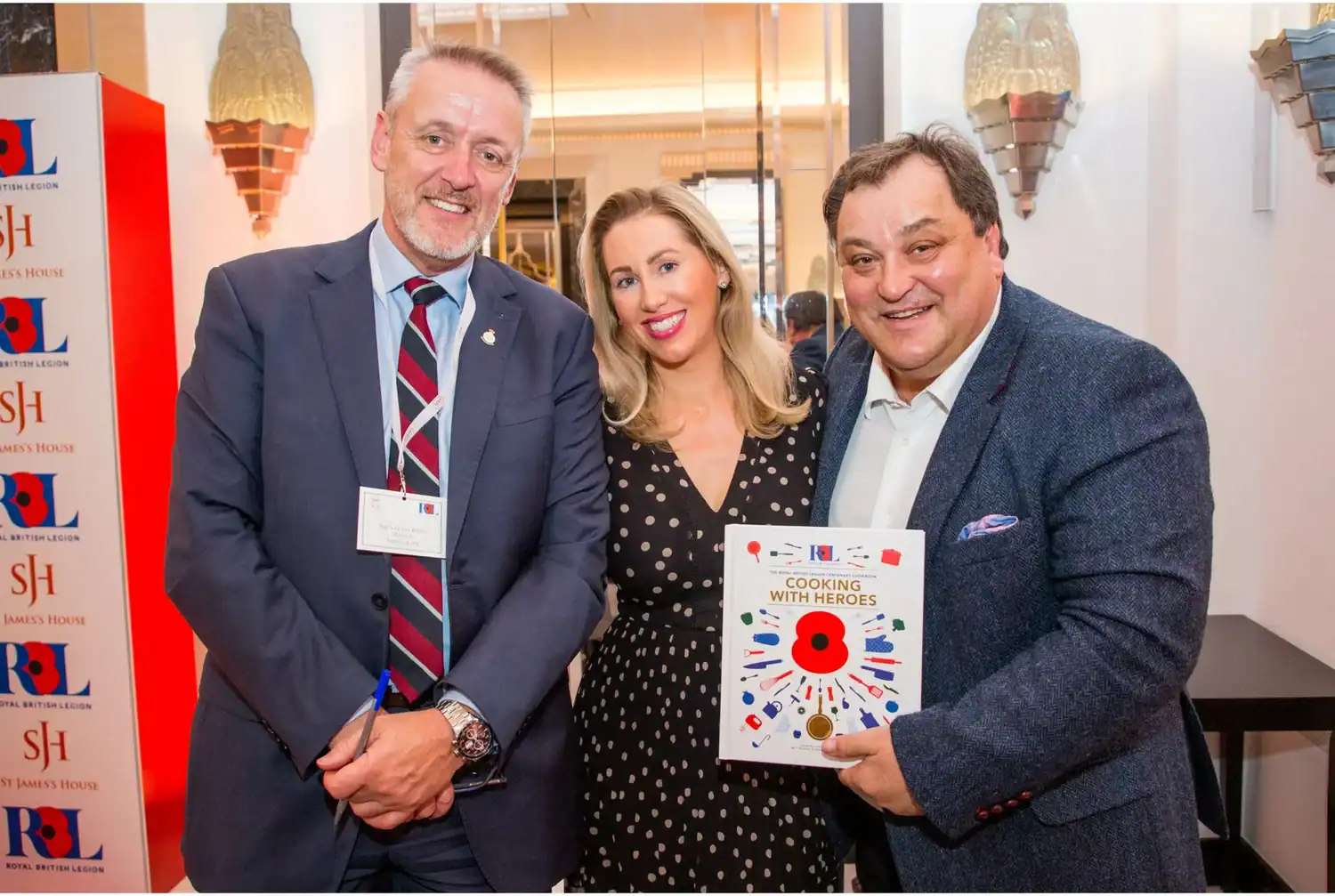 Three people posing indoors. The middle person smiles with two others. One proudly holds the book "Cooking with Heroes," blending the art of culinary tales and heroic deeds. The background features logos and decorative lights, adding a touch of glamour to this momentous occasion.