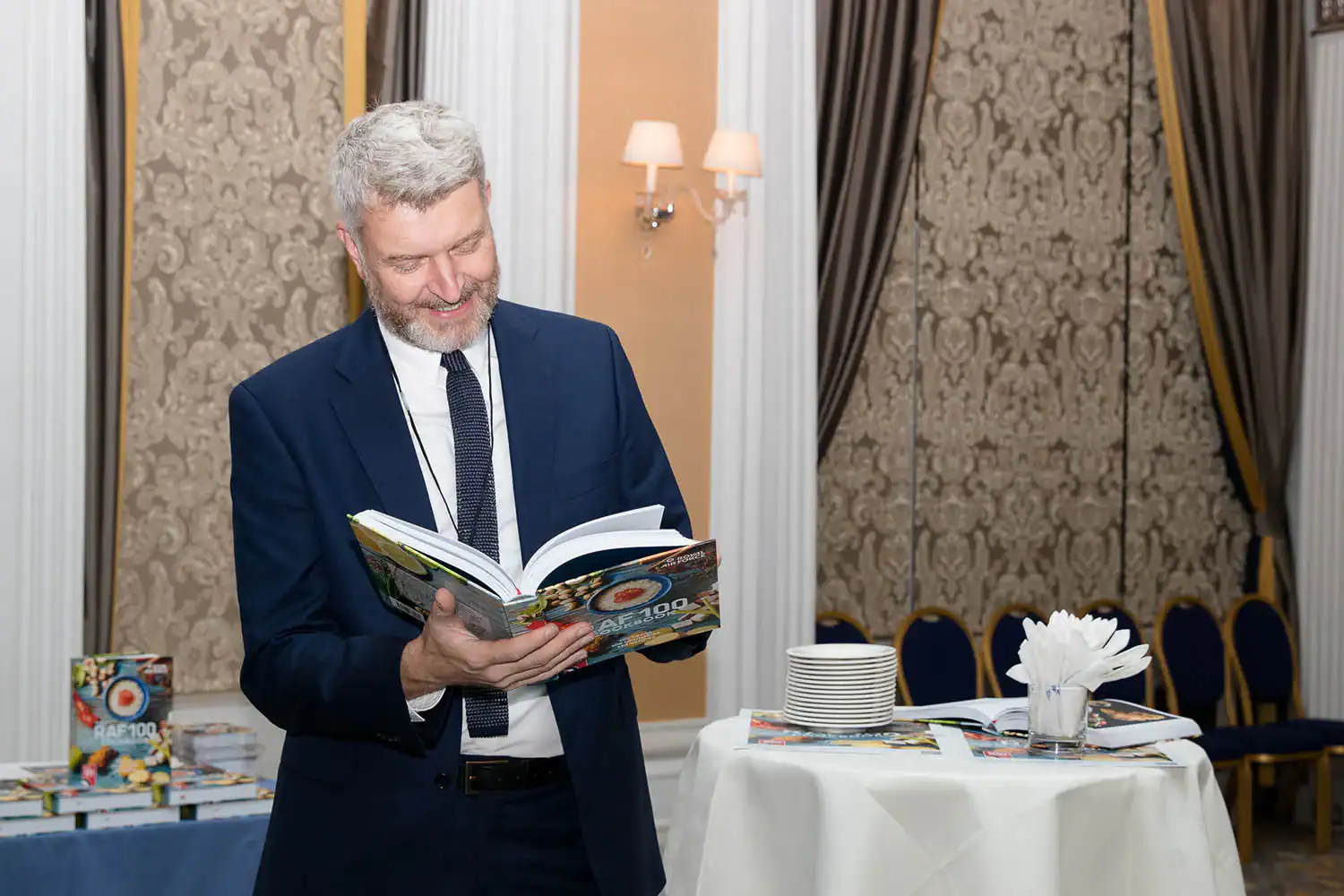 A man in a suit reads a large cookbook at a formal event, standing beside a table with stacked dishes and napkins. Several copies of the same book are visible in the background, perhaps part of the RAF 100 celebration's culinary collection.