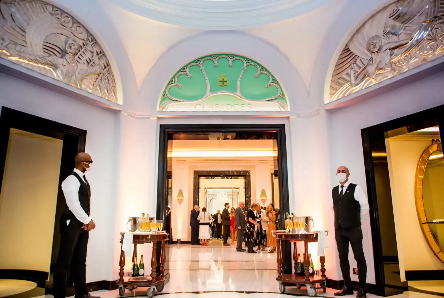Elegant hotel entrance with art deco design, two uniformed staff standing by like heroes of hospitality, and guests mingling in the background under ornate archways.