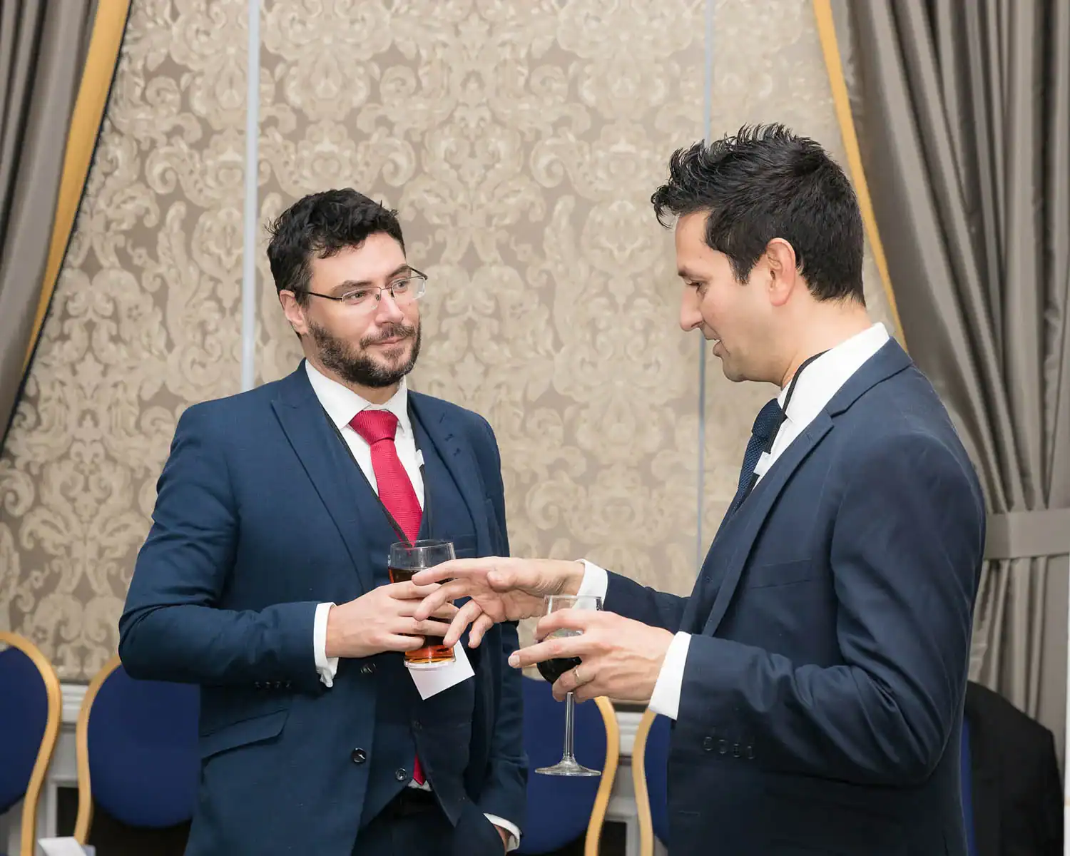 Two men in formal suits are conversing in a decorated room, both holding drinks. A copy of the RAF 100 Cookbook rests on a nearby table, adding an unexpected touch to the elegant setting.