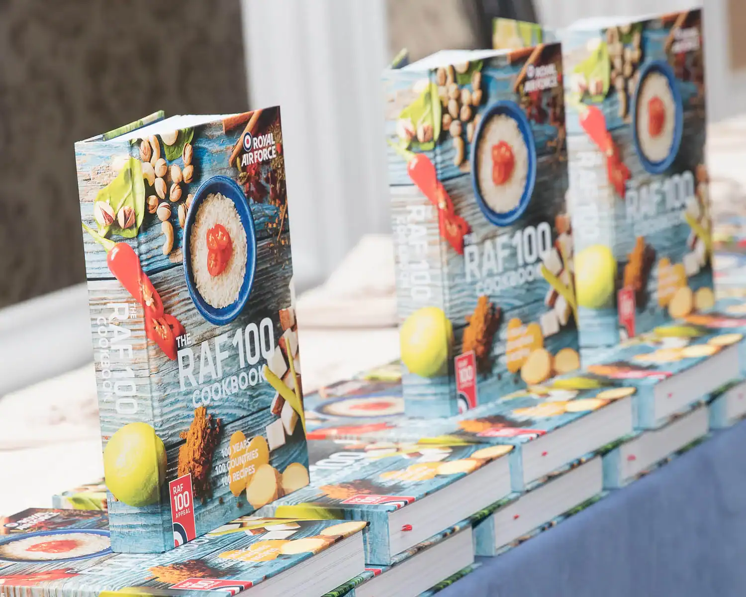 A stack of "The RAF 100 Cookbook" graces the table, showcasing its colorful cover adorned with a variety of tempting dishes alongside the distinguished Royal Air Force emblem.