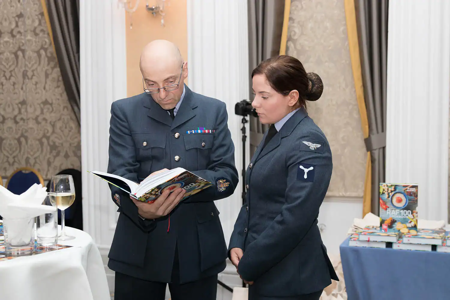 Two people in military uniforms stand indoors. The person on the left is flipping through an open RAF 100 Cookbook, while the person on the right observes with interest. A table with wine and more books is beside them.