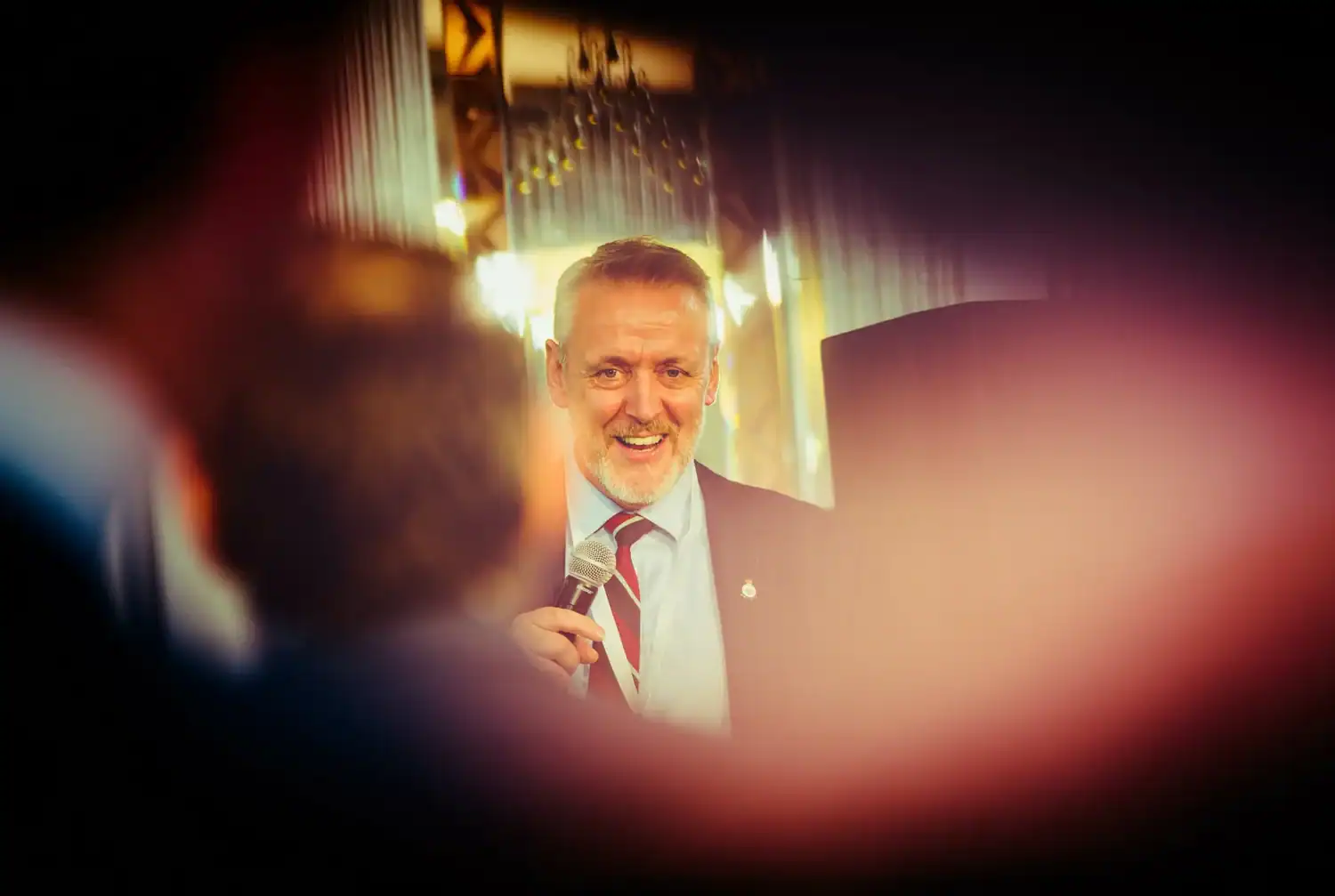A man in a suit and tie holds a microphone, speaking indoors with blurred figures in the foreground, delivering an inspiring tribute to everyday heroes.