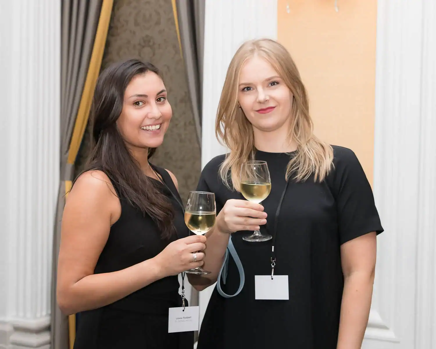 Two women in black dresses, perhaps capturing the elegance of an era celebrated in RAF 100, hold glasses of white wine. Standing indoors with name tags, they exude a timeless grace amidst the event's lively backdrop.