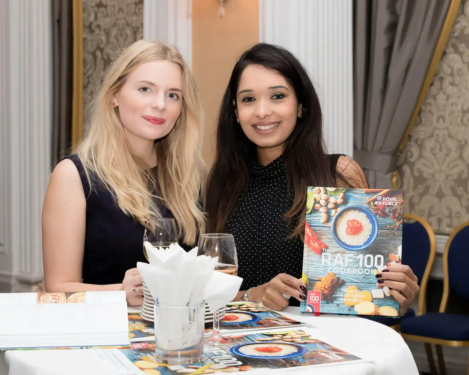 Two women are seated at a table, enjoying drinks while immersed in the "RAF 100 Cookbook." Amidst ornate decor and a tall mirror, the table is scattered with more books and a napkin holder, creating an inviting atmosphere for culinary inspiration.