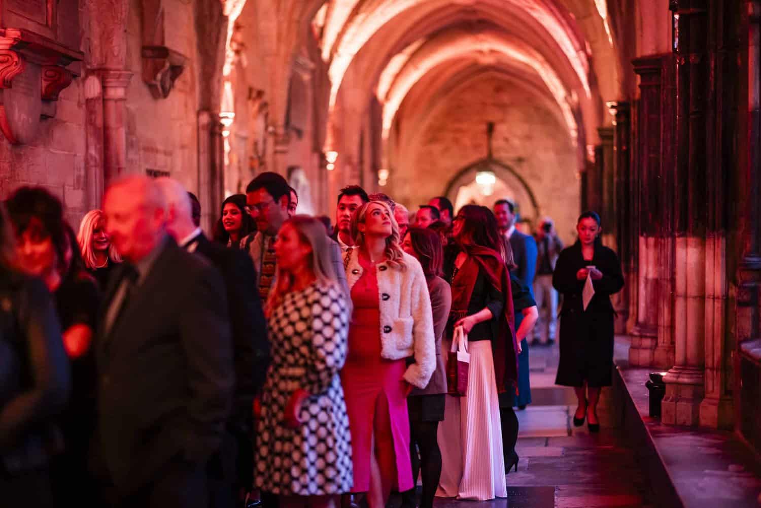 In a dimly lit, arched corridor bathed in red light, people gather to mark the Commonwealth's 75th Anniversary. Some are deep in conversation while others face forward, absorbing the historic ambiance.