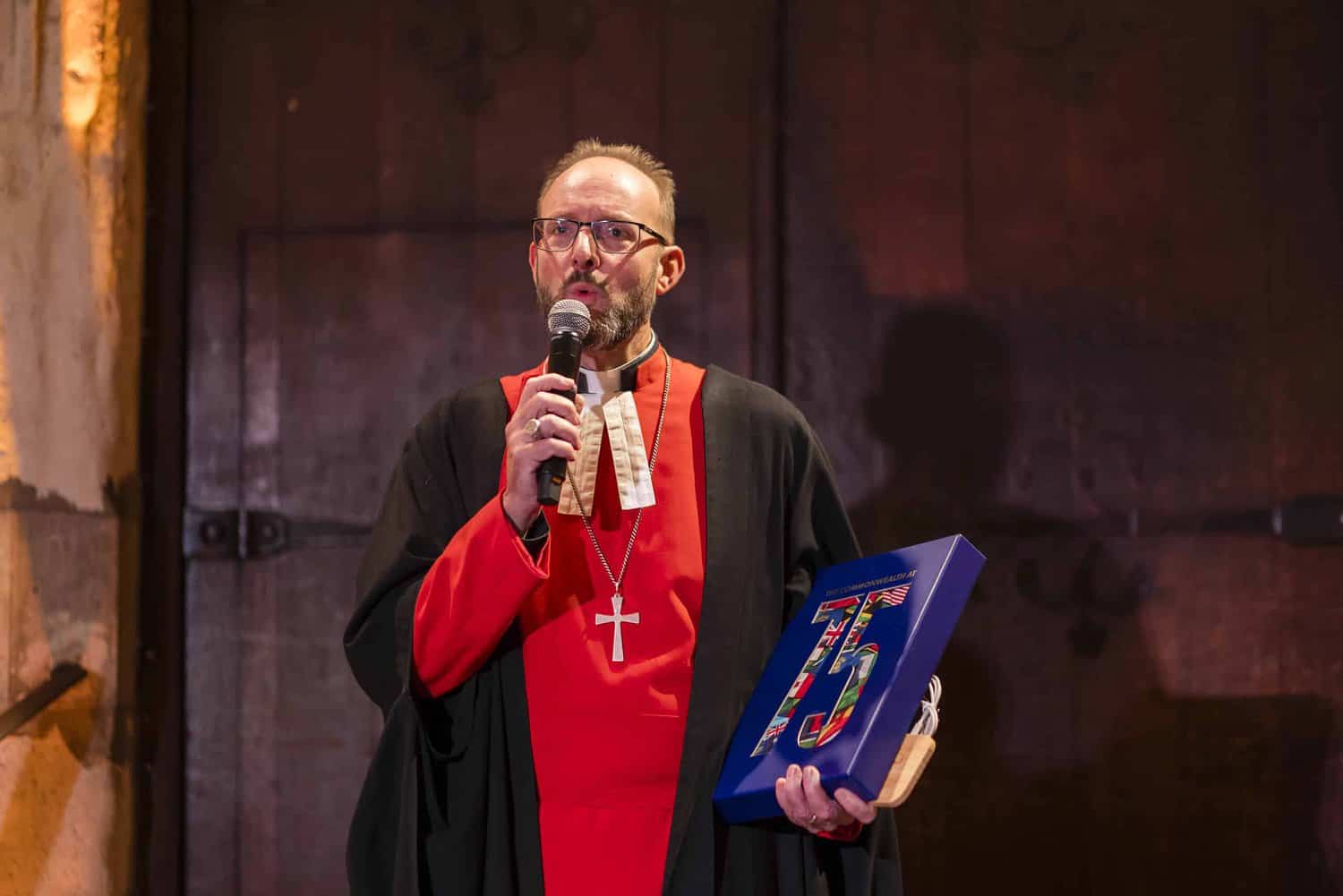 A person in a red and black robe with a cross necklace holds a microphone and a blue plaque with "75" on it, standing on stage, celebrating the 75th anniversary of Commonwealth history.