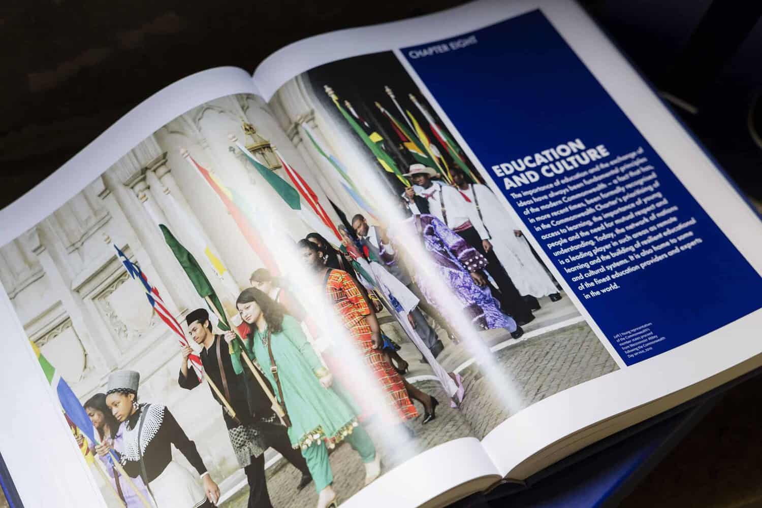An open book reveals a page with a photo of diverse people in cultural attire marching with flags, beneath the bold text "Education and Culture" on a blue background. This powerful image celebrates the 75th anniversary, marking a milestone for the Commonwealth's shared heritage.