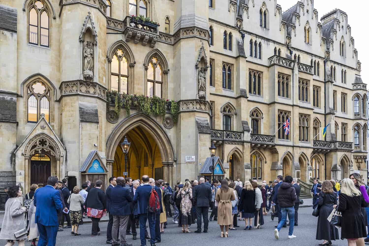 A group of people gathered outside a historic building with gothic architecture, adorned with numerous flags, in celebration of the Commonwealth's 75周年.