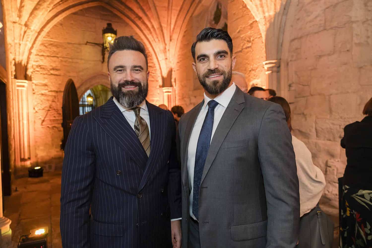 Two men in suits stand smiling in a stone archway room with warm lighting, celebrating Commonwealth history on its 75th anniversary.