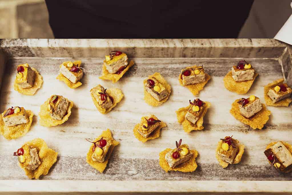 Wooden tray with appetizers, each consisting of a cracker topped with a small piece of pâté, a dollop of sauce, and garnish, crafted to honor the Commonwealth's rich history.