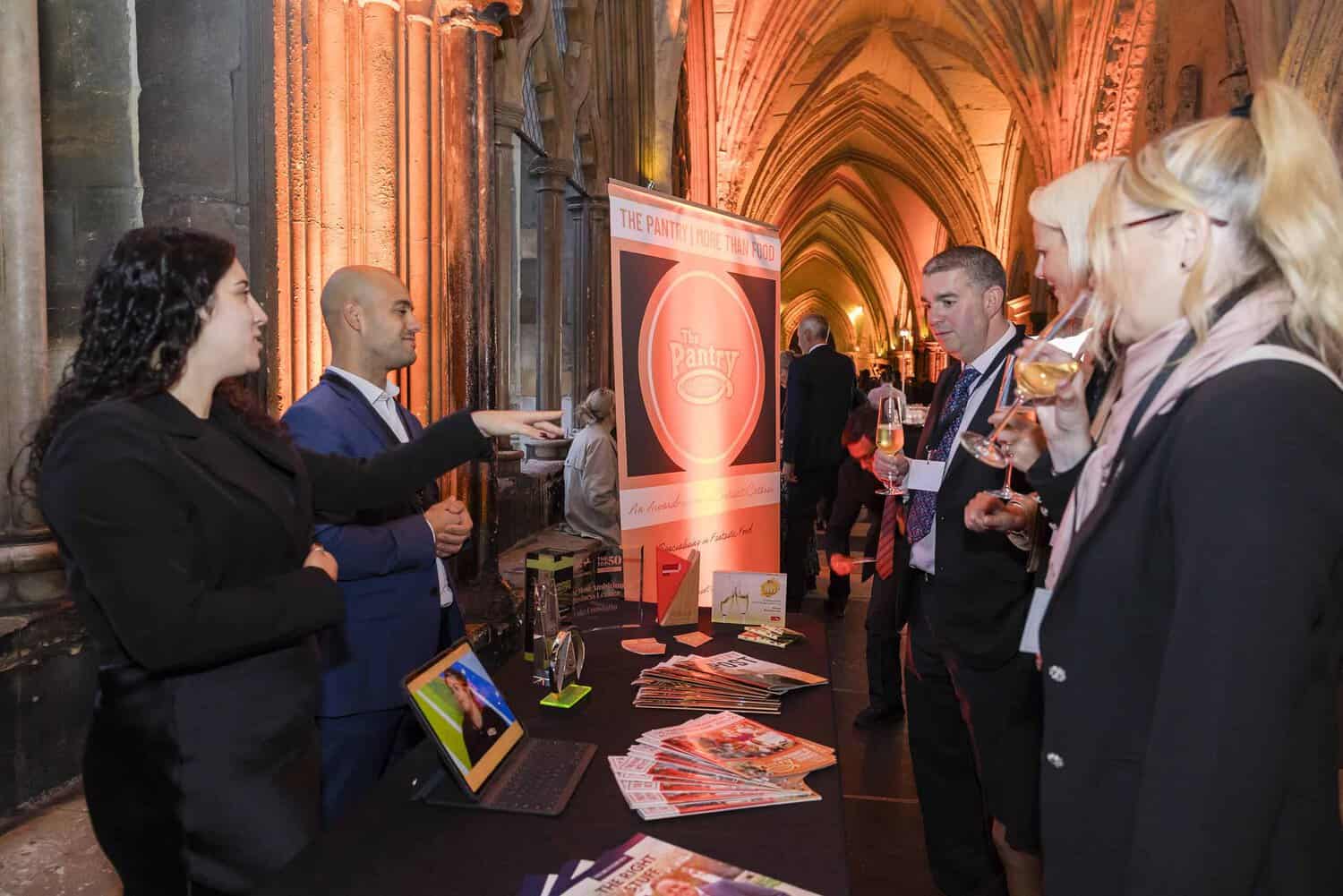 People gather around a table with brochures and a laptop beneath a banner reading "The Pantry." Celebrating the 75th anniversary of this esteemed Commonwealth event, they converse and hold drinks inside a building adorned with elegant arched architecture.