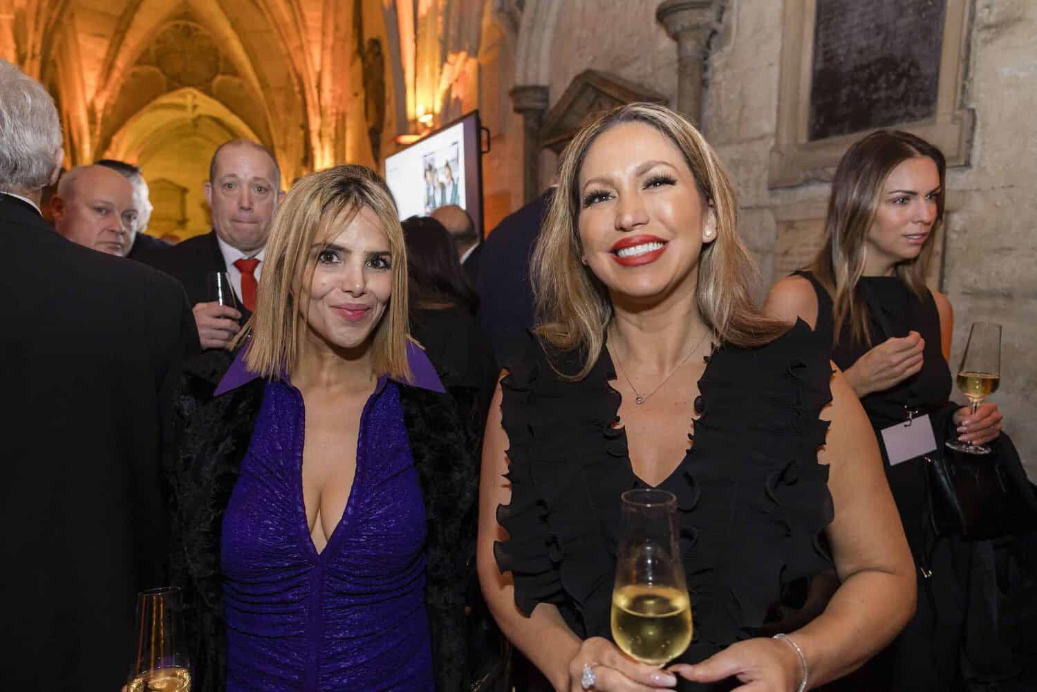 Two women smile and hold wine glasses at a formal event celebrating the 75th anniversary of the Commonwealth, set in a grand hall with stone arches, surrounded by other attendees.
