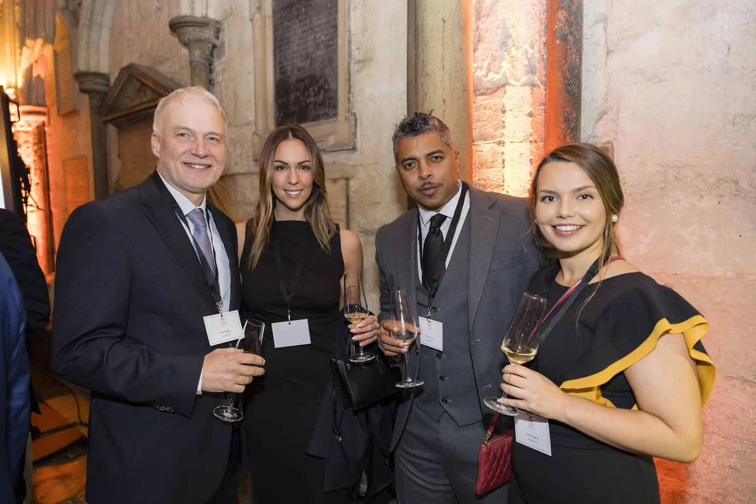 Four people in formal attire, each holding wine glasses, pose elegantly indoors at an event celebrating the 75th Anniversary of the Commonwealth.