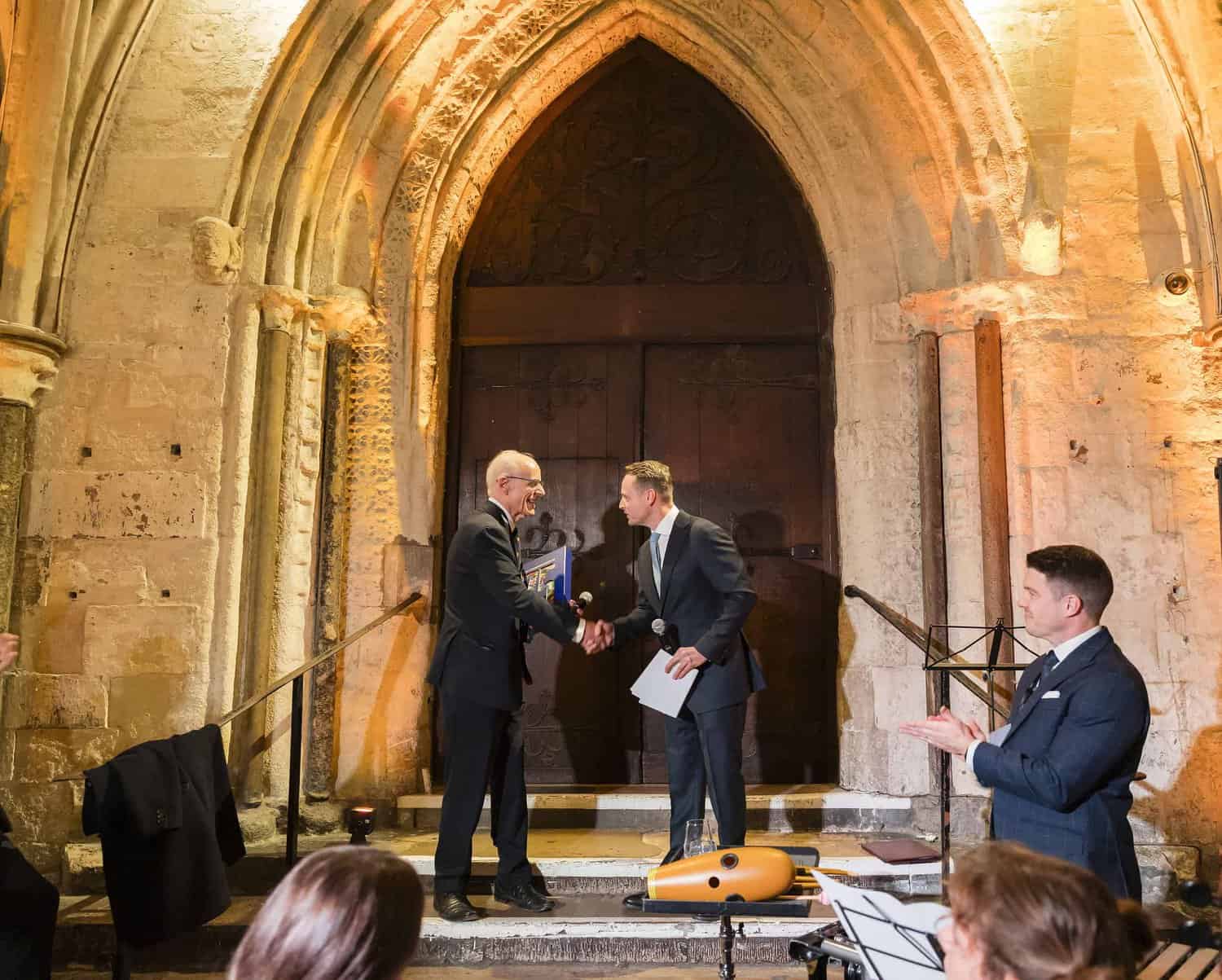 Two men in suits shake hands on the steps of an ornate stone doorway, marking a momentous agreement. Nearby, another man takes notes, capturing the day's details. In the foreground, a musical instrument and sheet music hint at upcoming Commonwealth celebrations for the 75th anniversary.