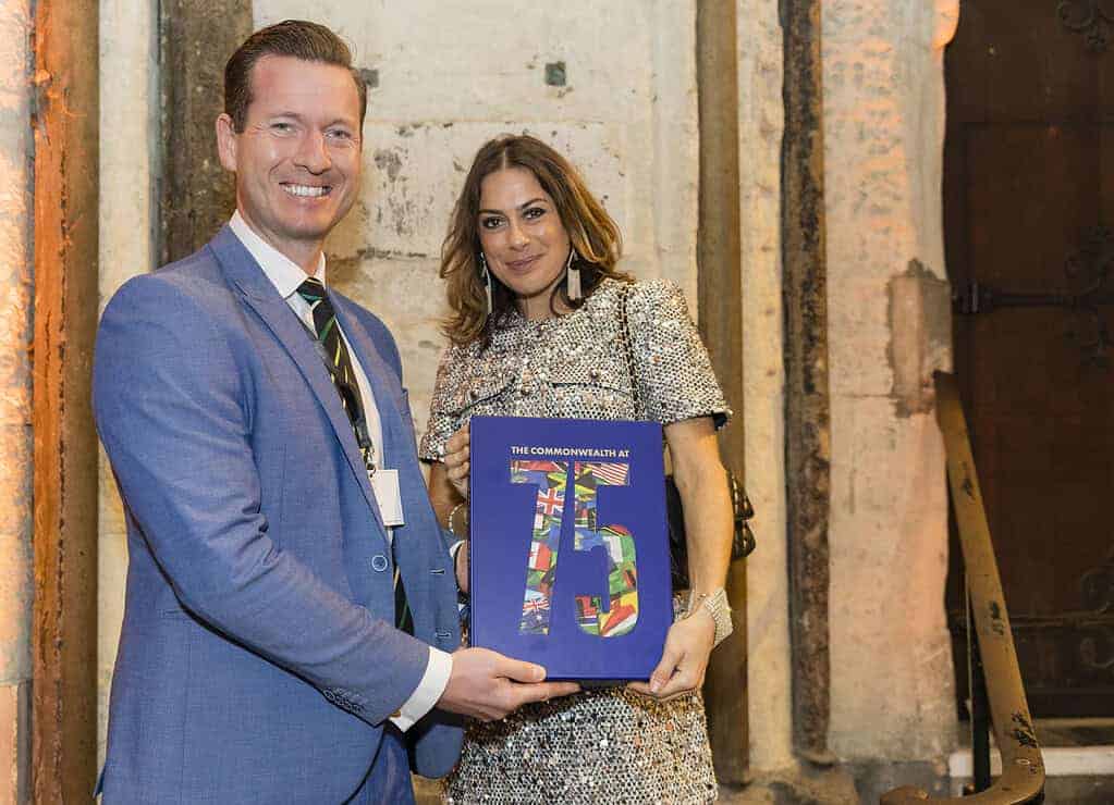 Two people smiling and holding a book titled "The Commonwealth at 75" celebrate the 75th Anniversary of the Commonwealth. The man, in a blue suit, and the woman, in a patterned dress, stand proudly before a worn stone doorway.