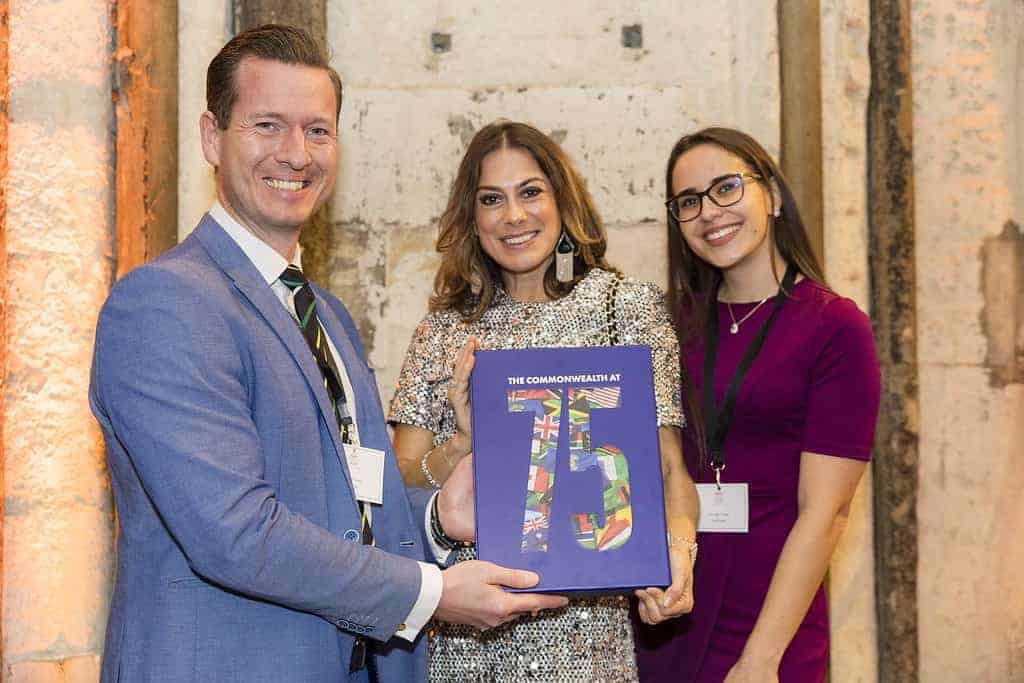 Three people smiling and holding a book titled "THE COMMONWEALTH AT 15" with a colorful cover, marking milestones achieved over the years, stand indoors against a textured wall.