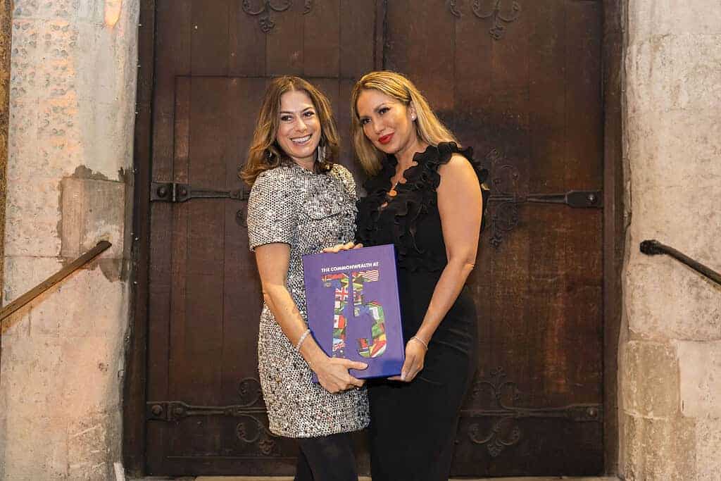 Two women stand together smiling, holding a colorful book titled "The Commonwealth at 70" in front of a wooden door, celebrating the global community's rich history ahead of its 75th anniversary.