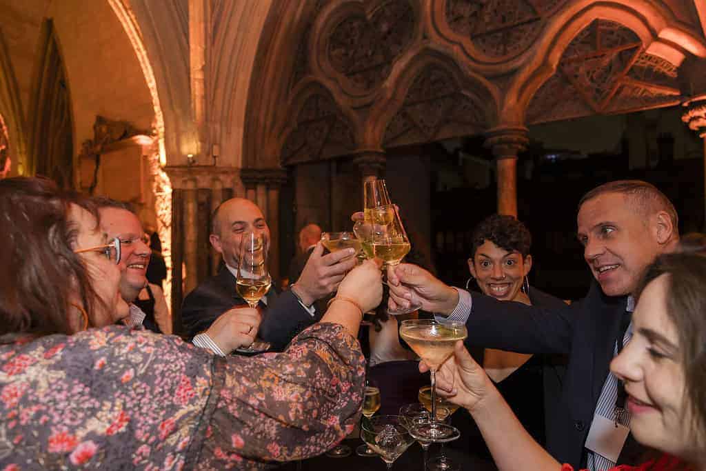 In an ornate, arched indoor setting, a group of people raises their glasses in a toast, celebrating the Commonwealth's 75th anniversary.