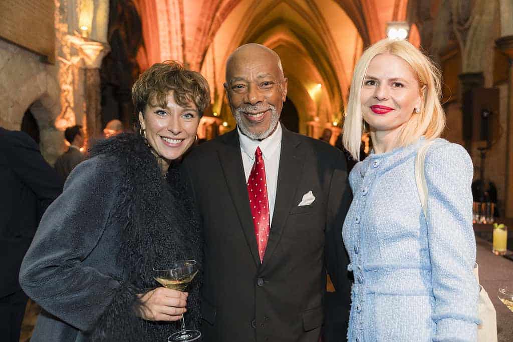Three people smiling at a formal event in a historic building with arches, celebrating the Commonwealth's 75th Anniversary. One holds a drink, capturing the essence of history in a single moment.
