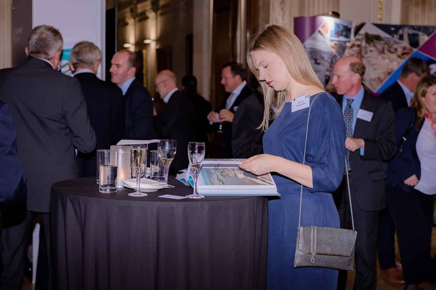 A woman in a blue dress reads a world classic at a cocktail table during a formal event, shaping her thoughts as guests converse in the background.