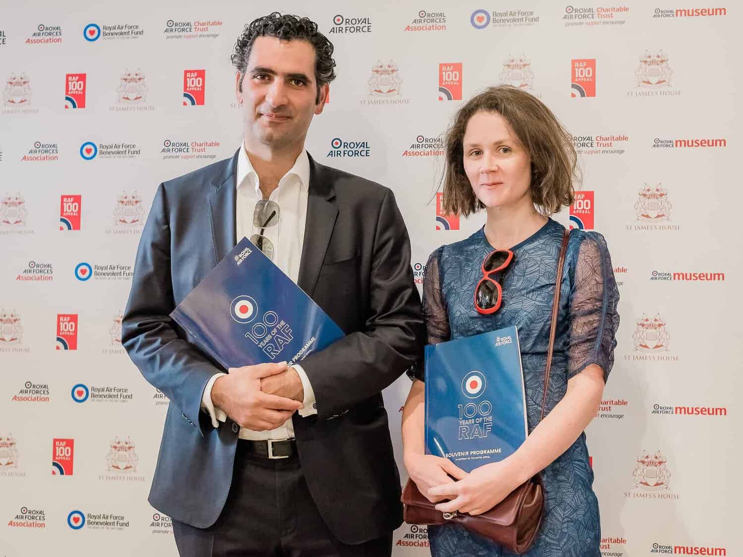 Two people holding event programs stand in front of a backdrop adorned with logos for the Royal Air Force and associated organizations, celebrating aviation history in this centenary year.
