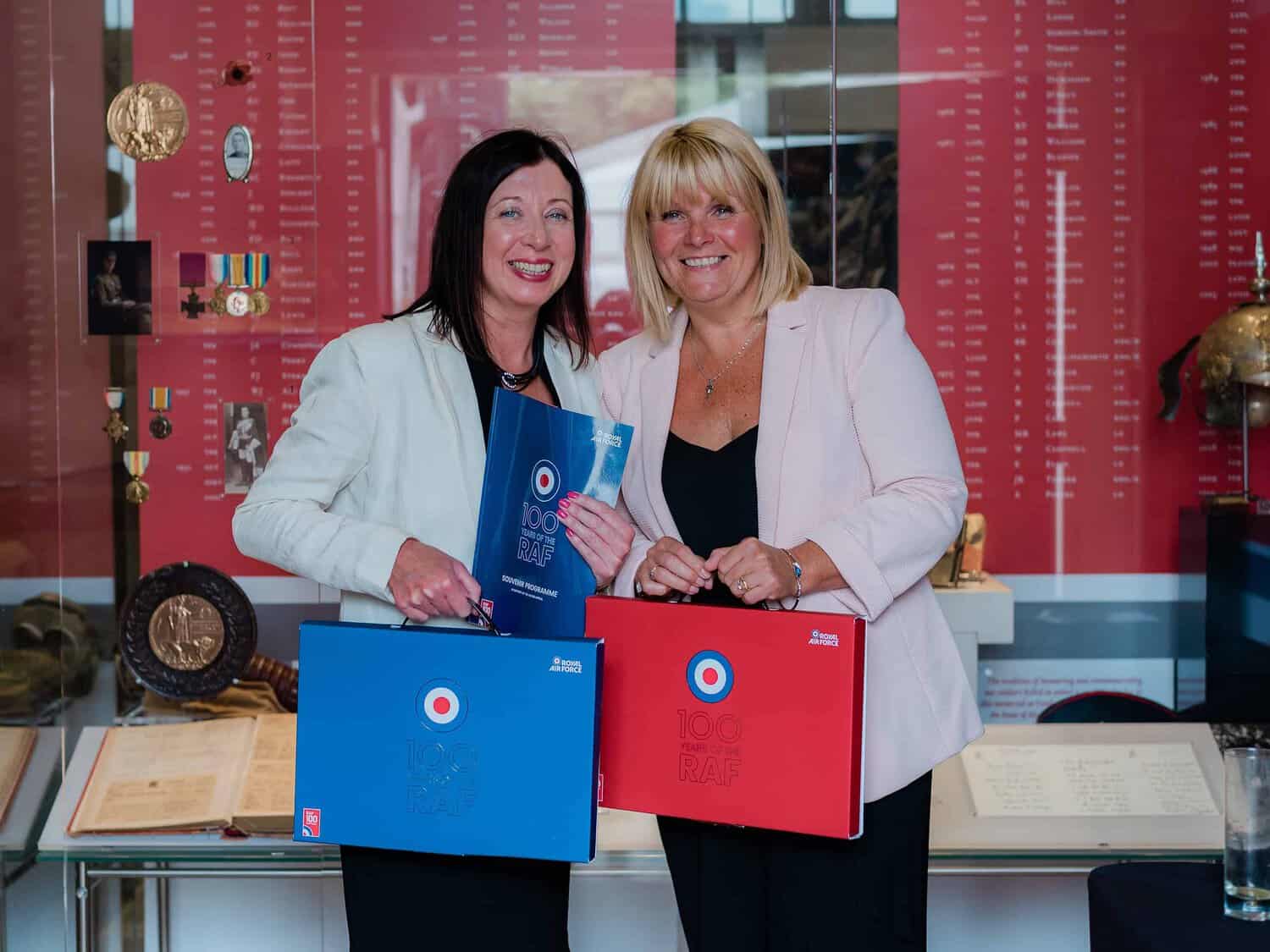 Two women stand side by side, each holding folders with "100 RAF" logos. They are smiling and dressed in business attire, celebrating 100 years of the Royal Air Force, with a display of medals and historical items in the background.