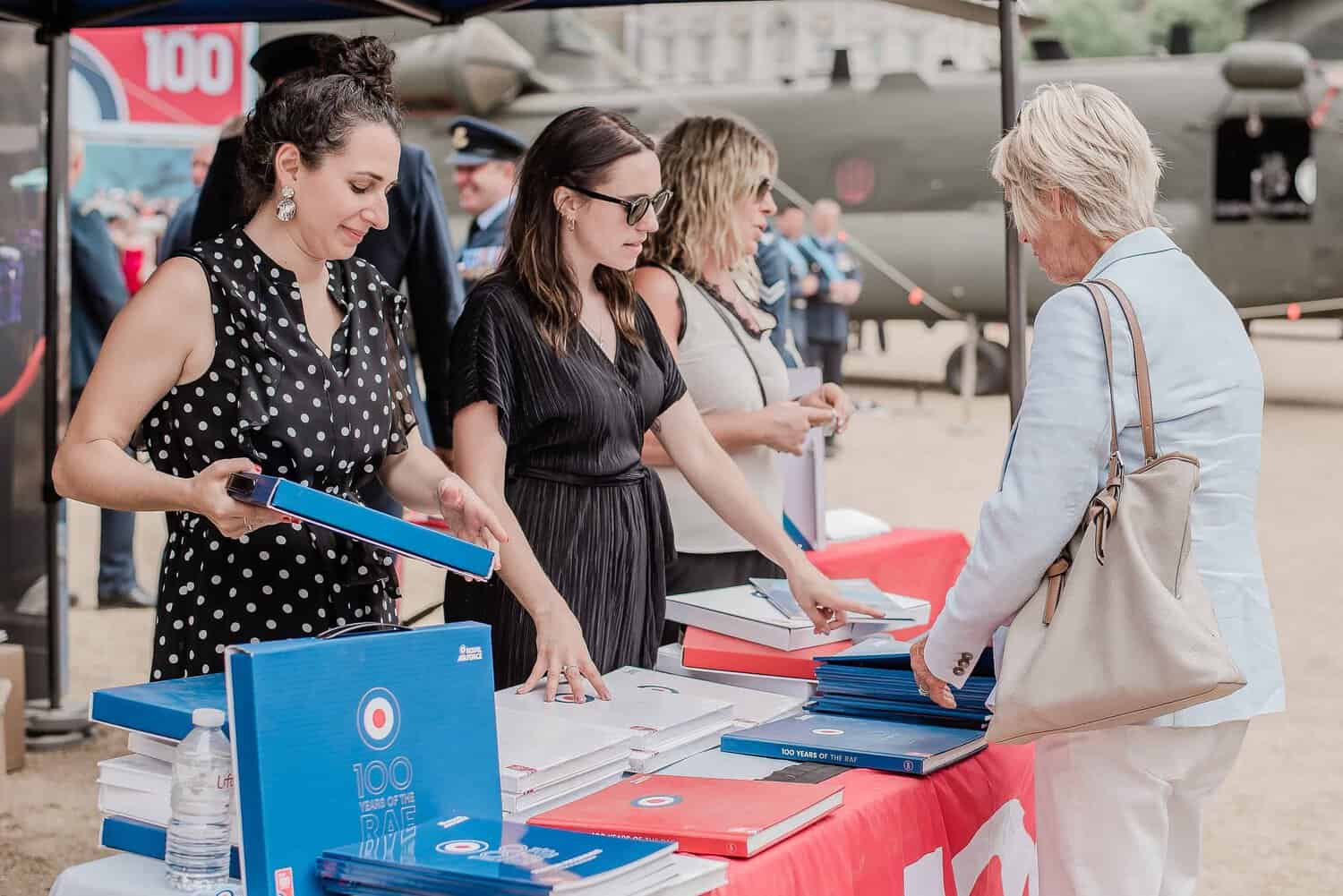 People at a table distribute books next to a military airplane display, celebrating 100 years of the Royal Air Force.