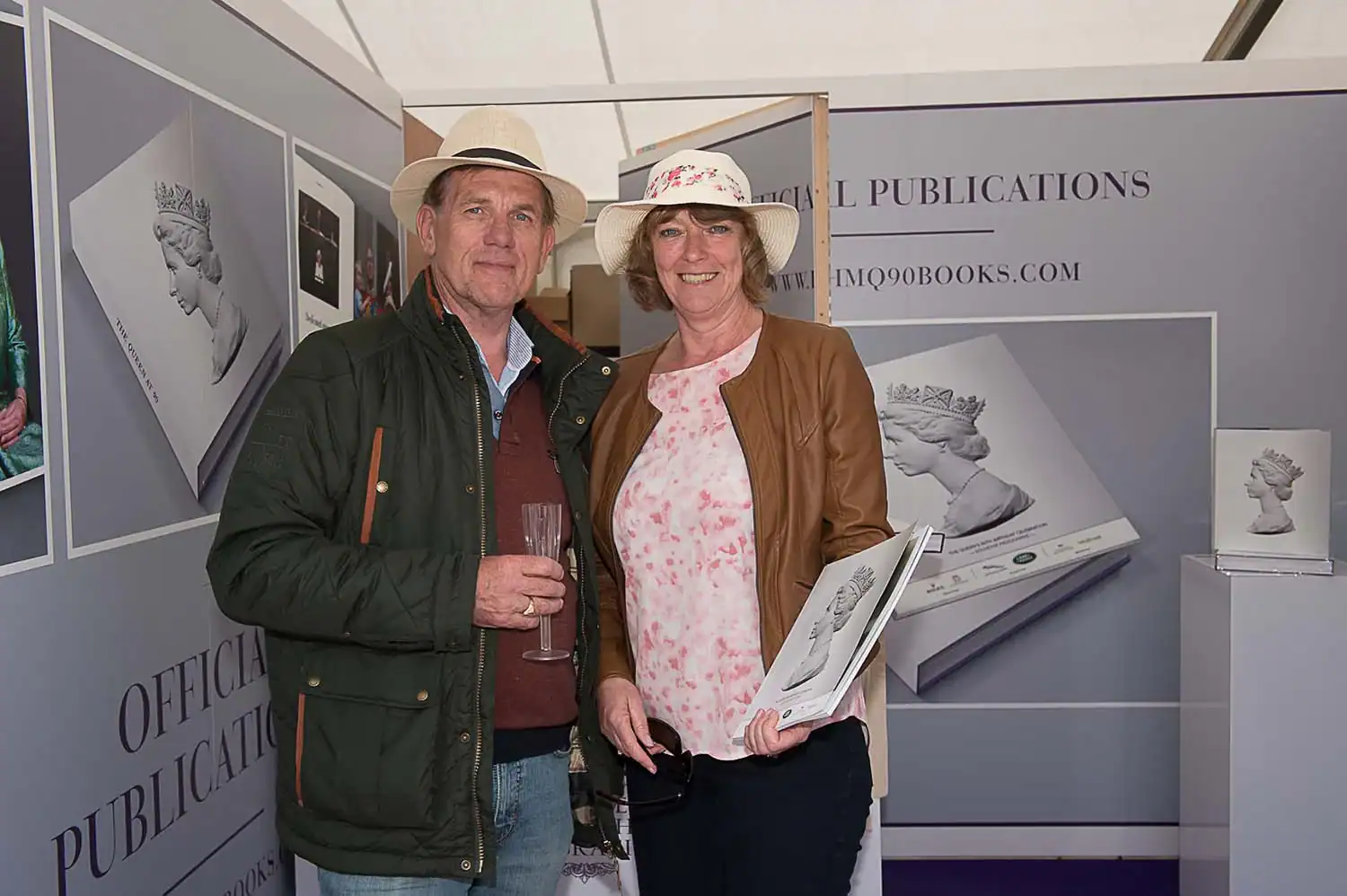 A man and woman wearing hats stand indoors holding a book about the Queen. They are in a booth displaying official publications, reminiscent of scenes from a fascinating documentary.