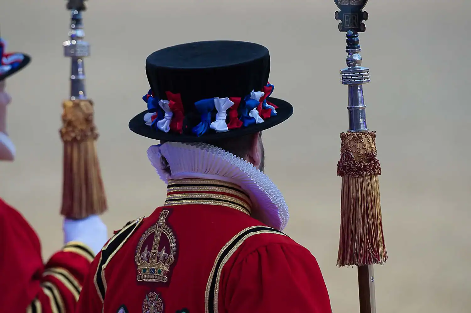A ceremonial guard in ornate red attire and a black hat with red, white, and blue ribbons stands proudly holding a staff with a decorative tassel, marking the Queen's 90th Birthday during the Royal Celebration.