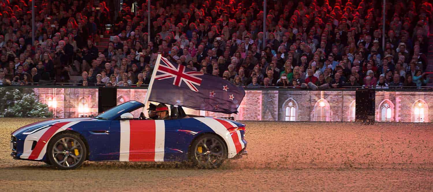 A sports car adorned with a Union Jack speeds across the arena surface, proudly displaying a large New Zealand flag. The scene unfolds before an audience of thousands, paying tribute with a nod to heritage and royalty.