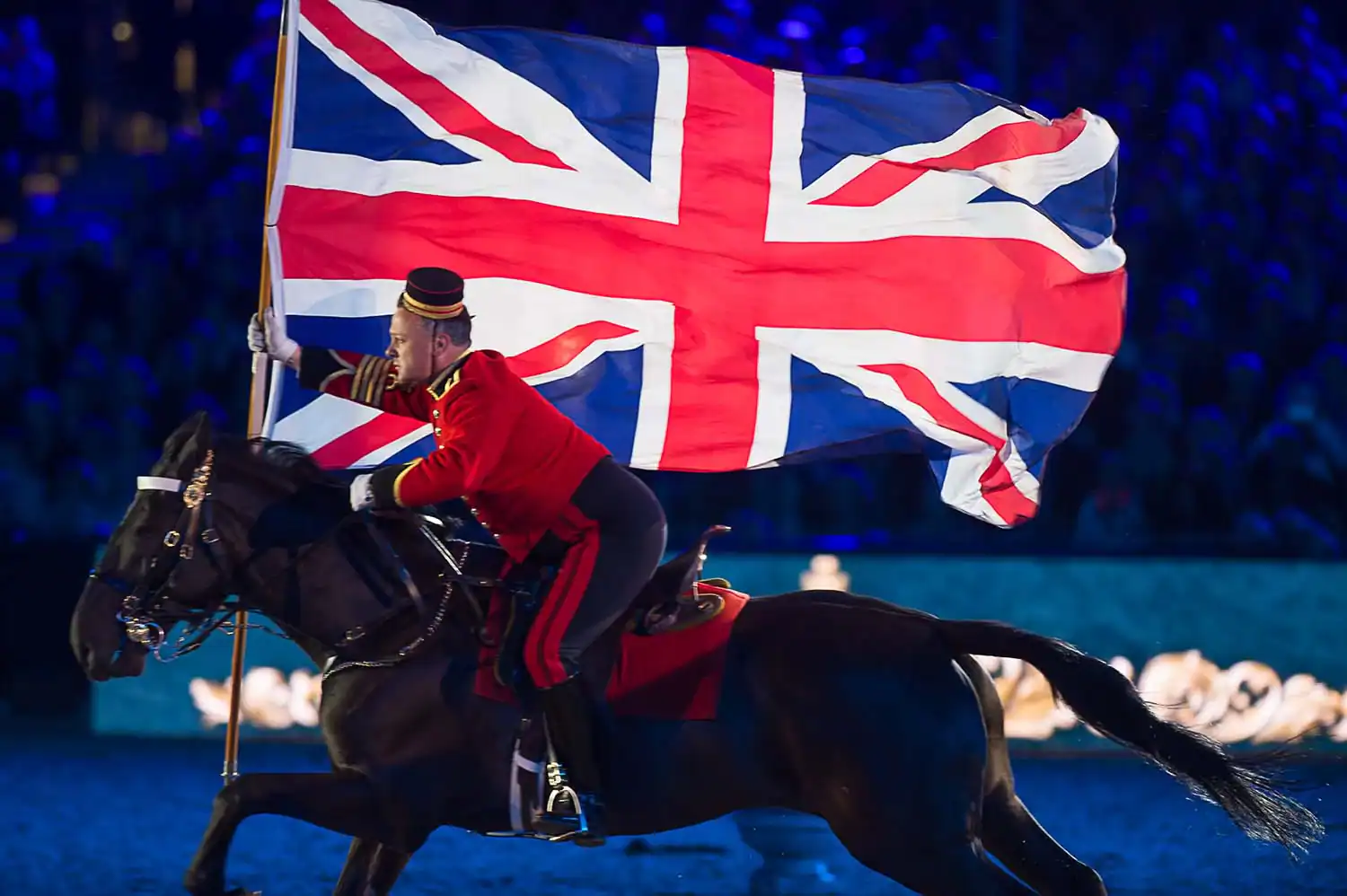 A person in a red uniform rides a horse while holding a large Union Jack flag against a dark background, reminiscent of the celebrations for Queen Elizabeth II's 90th birthday.