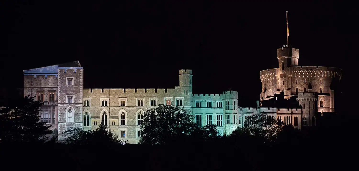 A large, illuminated castle at night with trees in the foreground and a flag atop a prominent round tower, echoing celebrations reminiscent of Queen at 90.