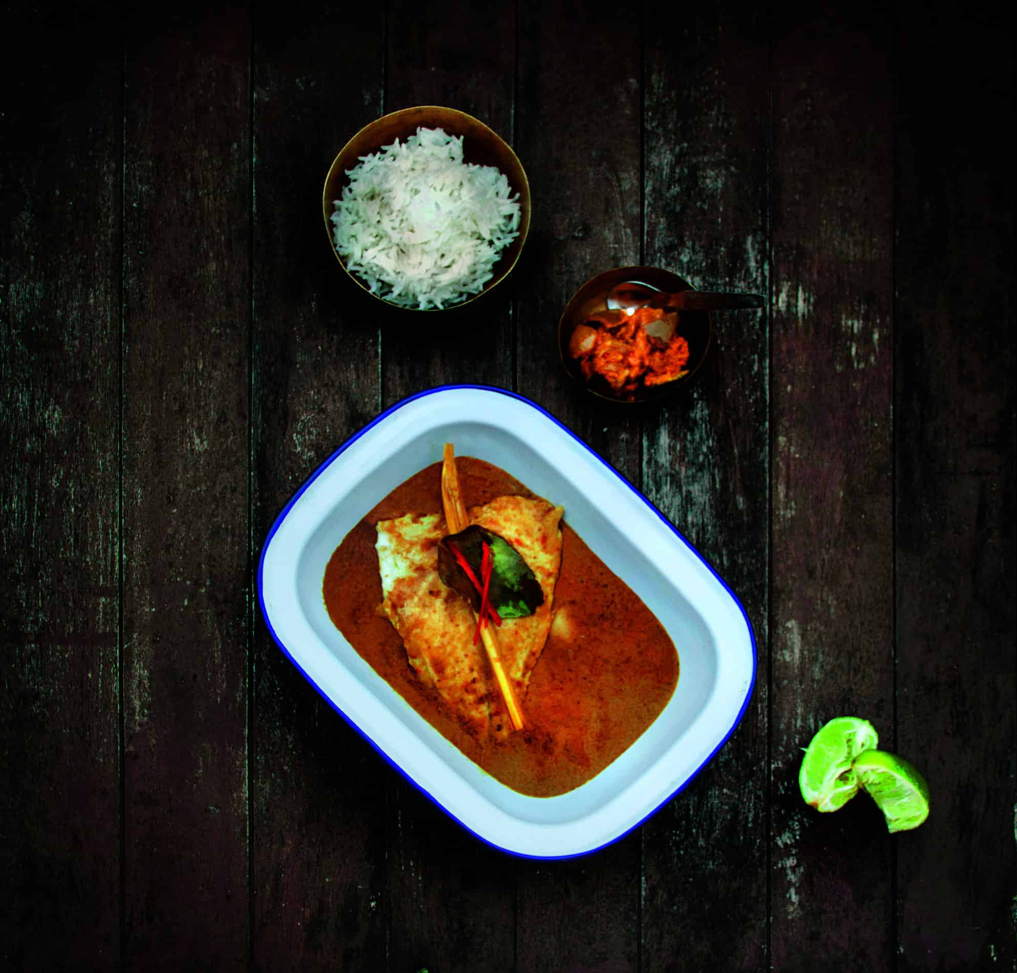 A dish with fish curry, inspired by the RAF 100 Cookbook, is elegantly presented in a rectangular bowl. It comes with a bowl of white rice and pickles. Lime wedges rest on the wooden table, adding a zesty touch to this culinary tribute to tradition and innovation.