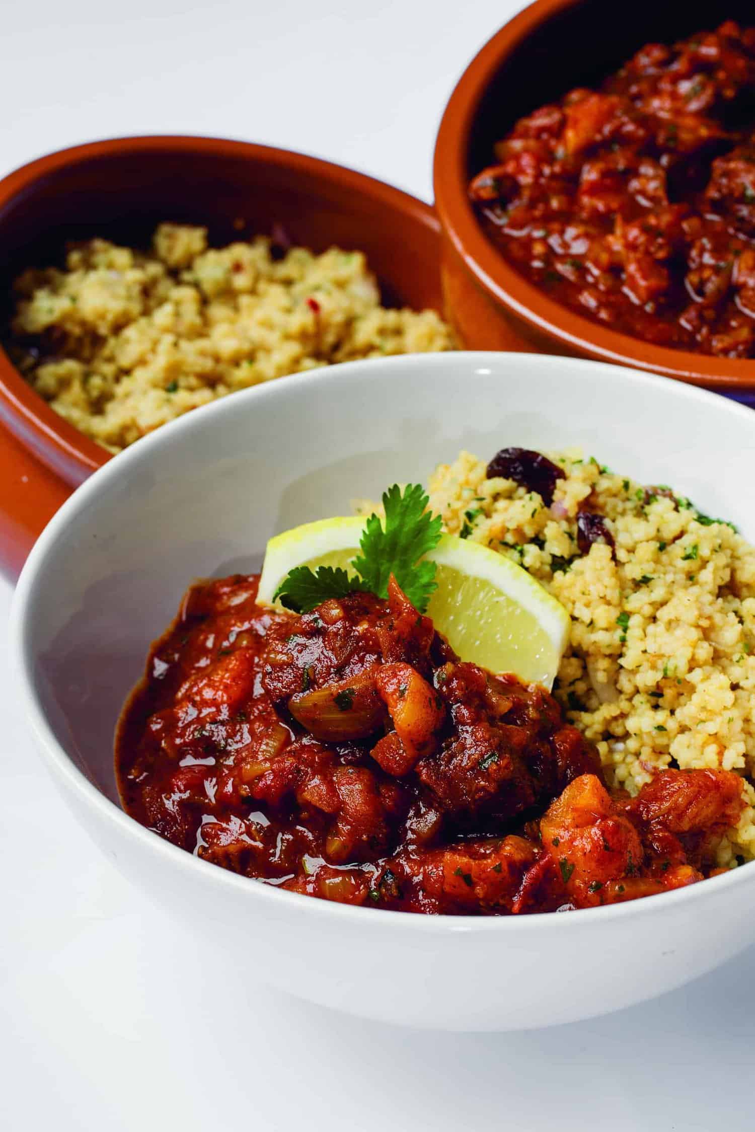 A bowl of chili with chunks of meat, garnished with a lime wedge and cilantro, served alongside couscous—a nod to the hearty recipes featured in the RAF 100 Cookbook. Two additional bowls of couscous and chili are in the background.