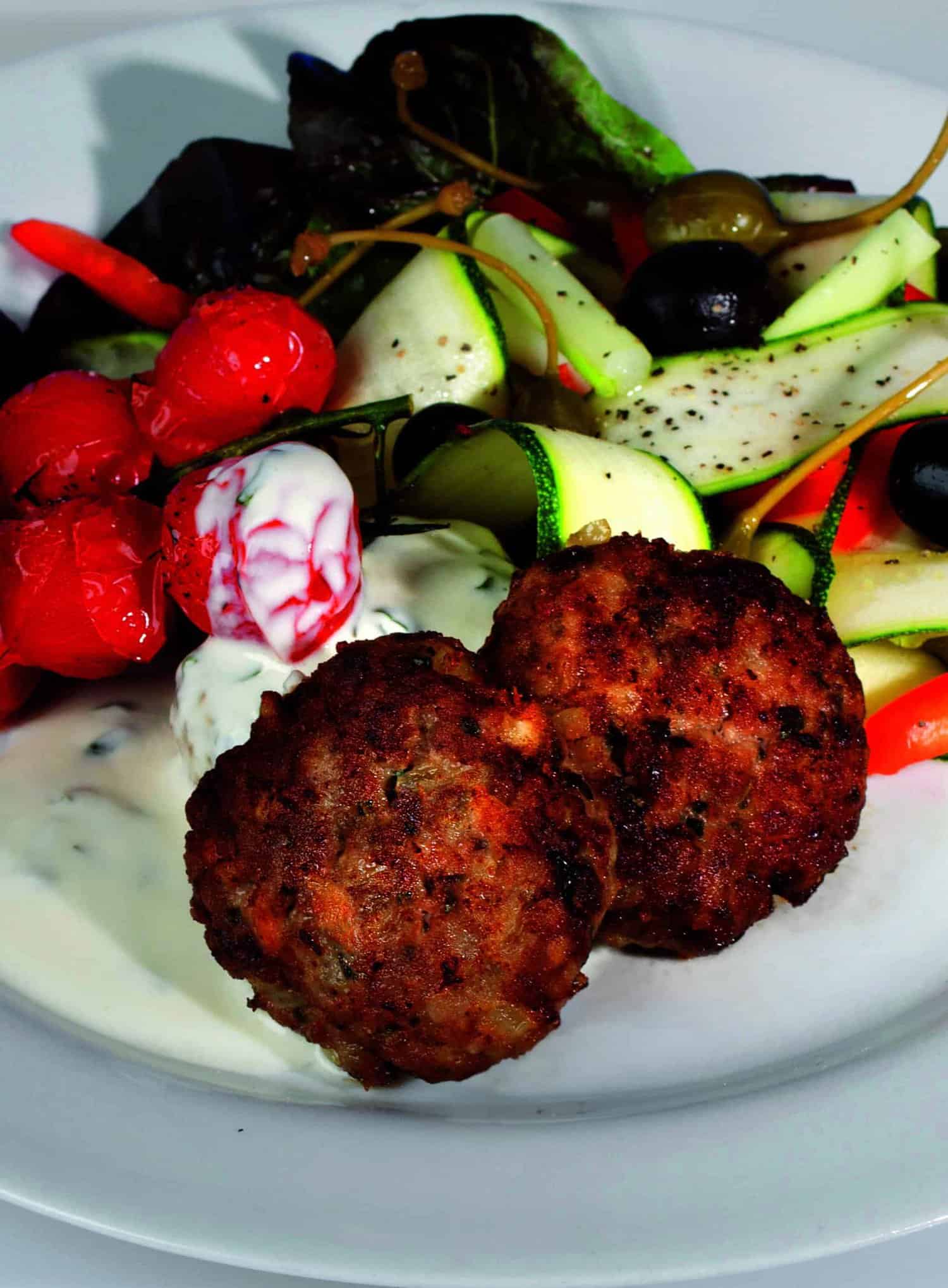 Two grilled meat patties on a plate with creamy sauce, garnished with raspberries, olives, zucchini strips, cherry peppers, and leafy greens—a delightful dish inspired by the RAF 100 Cookbook.