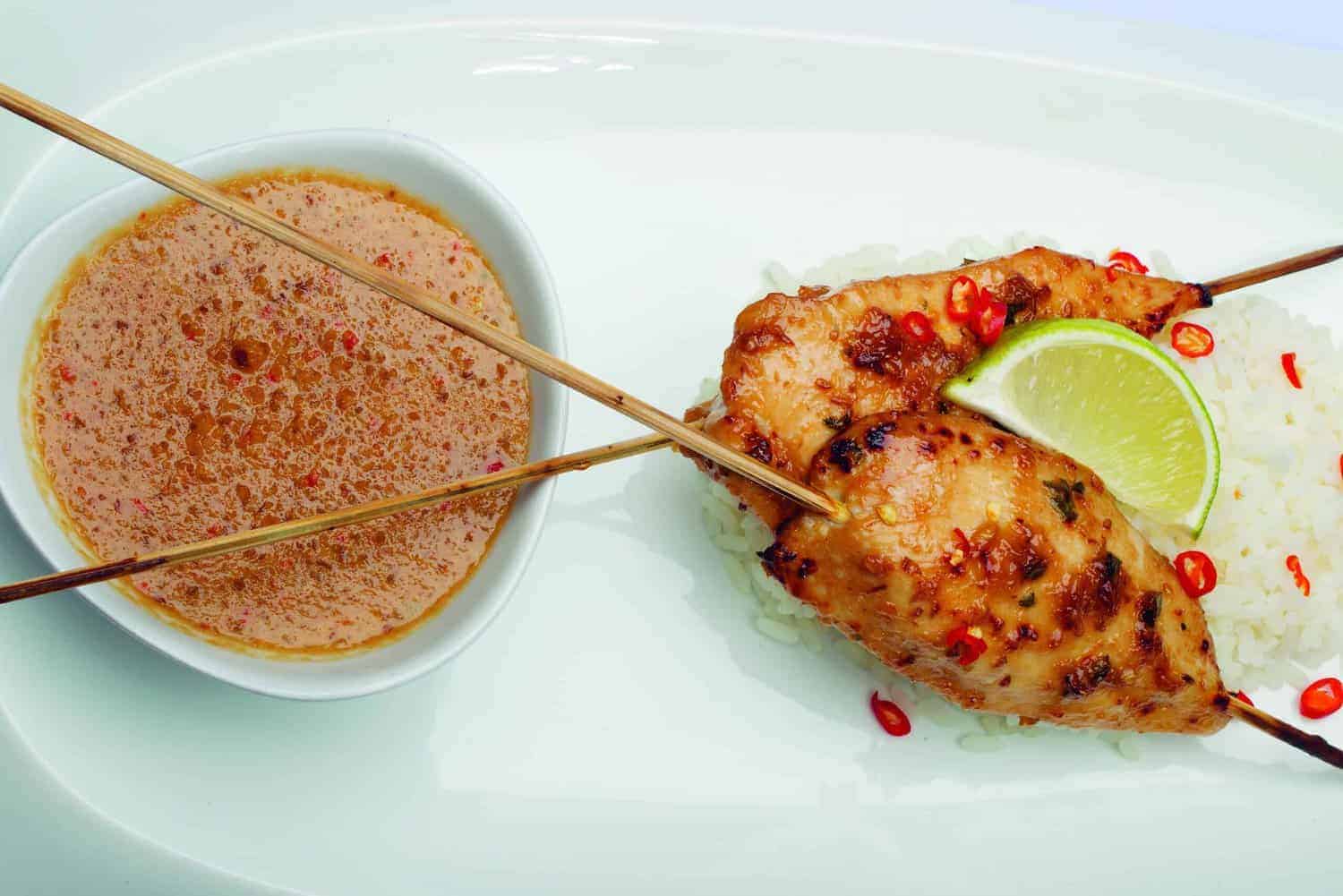 From the RAF 100 Cookbook, savor two grilled chicken skewers on rice with lime and chilies, elegantly served alongside a bowl of peanut sauce, celebrating culinary traditions with every bite.