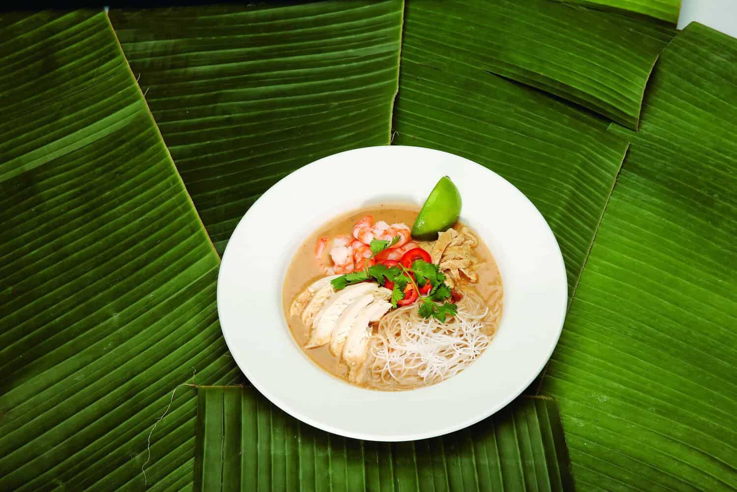 A bowl of noodle soup with sliced chicken, shrimp, red chili slices, and cilantro, garnished with a lime wedge, is set on a large banana leaf background—a delightful dish inspired by the RAF 100 Cookbook.
