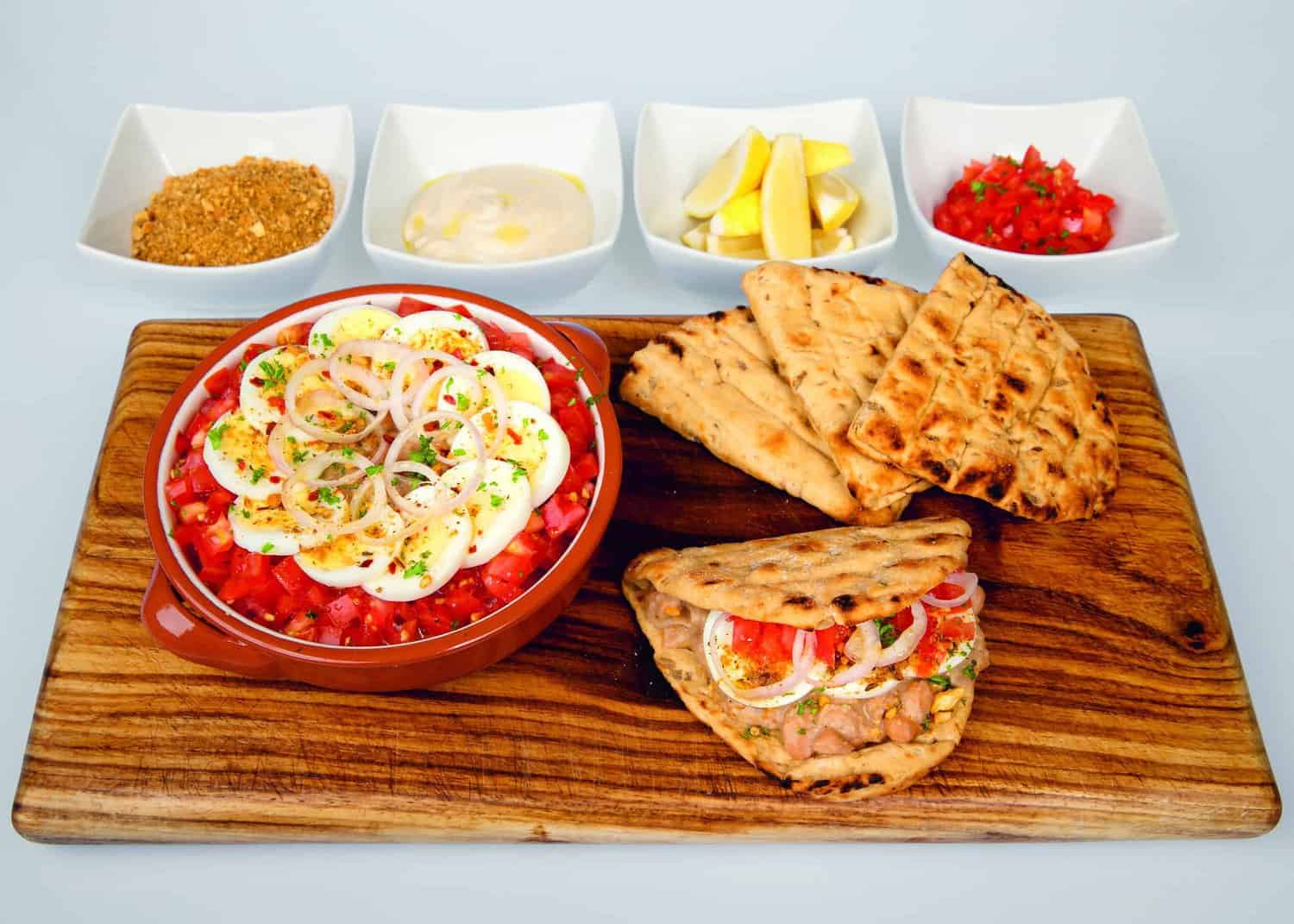 A cookbook-worthy bowl of salad with eggs and onions, accompanied by grilled flatbread on a wooden board. Behind are small dishes with spices, sauce, lemon wedges, and chopped tomatoes—a delight that could honor RAF 100 gatherings.