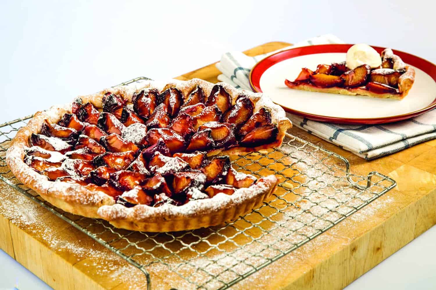 A baked tart topped with caramelized apple slices rests on a cooling rack, reminiscent of a classic recipe you'd find in an RAF 100 cookbook. A slice is served on a plate with a scoop of ice cream beside it.